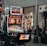 A collection of slot machines is situated inside a building with bright, colorful screens displaying various games and gaming themes. A person is seated nearby, seemingly relaxed with luggage beside them, and the atmosphere is lively with electronic displays.
